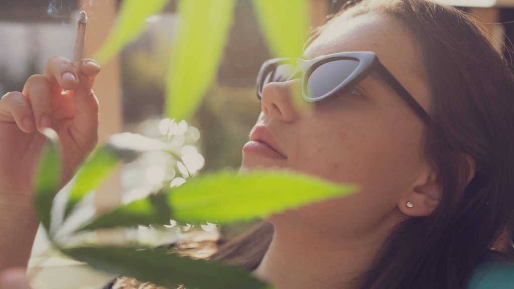 Frau mit Sonnenbrille raucht einen Joint, unscharfe Cannabispflanze im Vordergrund, Sonnenlicht im Hintergrund