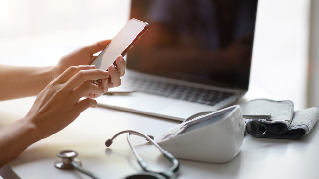 Frau mit Smartphone vor einem Laptop; auf dem Tisch liegen Blutdruckmessgerät und Stethoskop – Symbolbild für Telemedizin und medizinisches Cannabis auf Rezept.